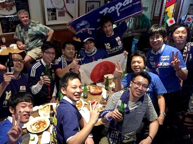 A group of sports fans are cheering for their national soccer team and one of them is wearing a shirt that says Japan.