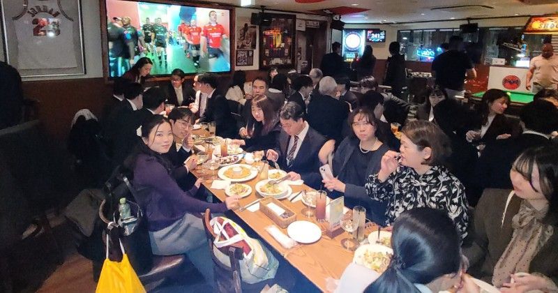 People eating and conversing in a crowded restaurant with a large screen showing sports.