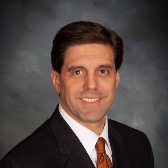 Man in suit smiling, with a brown tie, against a dark background.