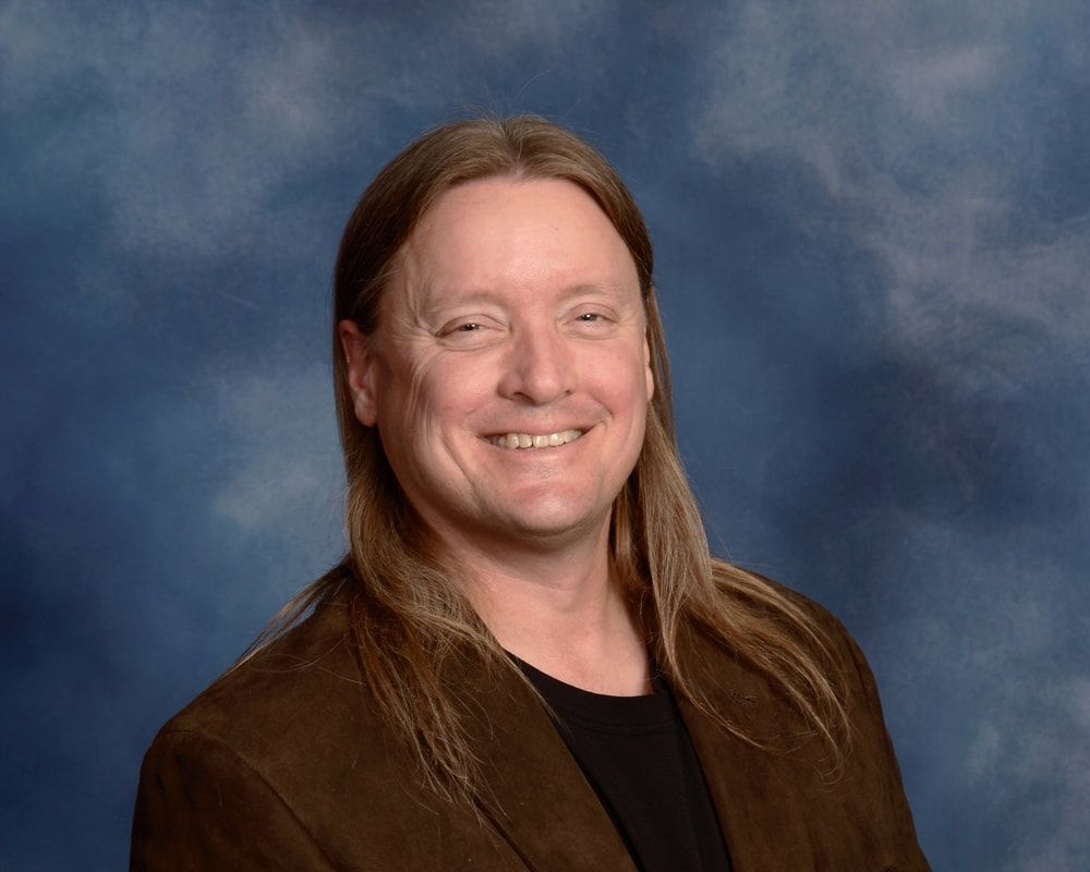 Man with long light brown hair smiles at the camera, wearing a brown jacket over a black shirt, blue background.