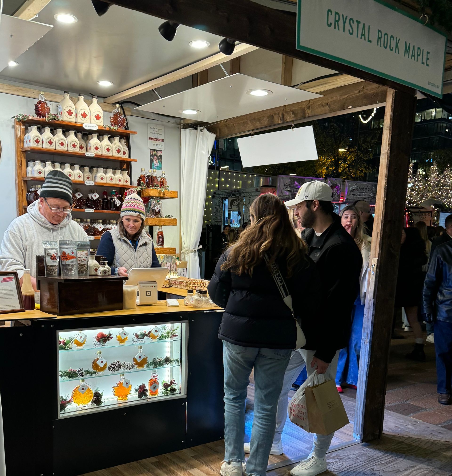 A maple syrup stand with two vendors, a couple shopping, bottles of syrup, and a sign that says 