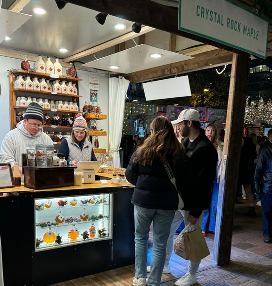 A maple syrup stand with two vendors, a couple shopping, bottles of syrup, and a sign that says