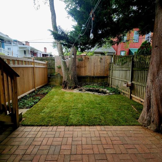 Backyard with brick patio, small lawn, wooden fence, trees, and buildings in background.