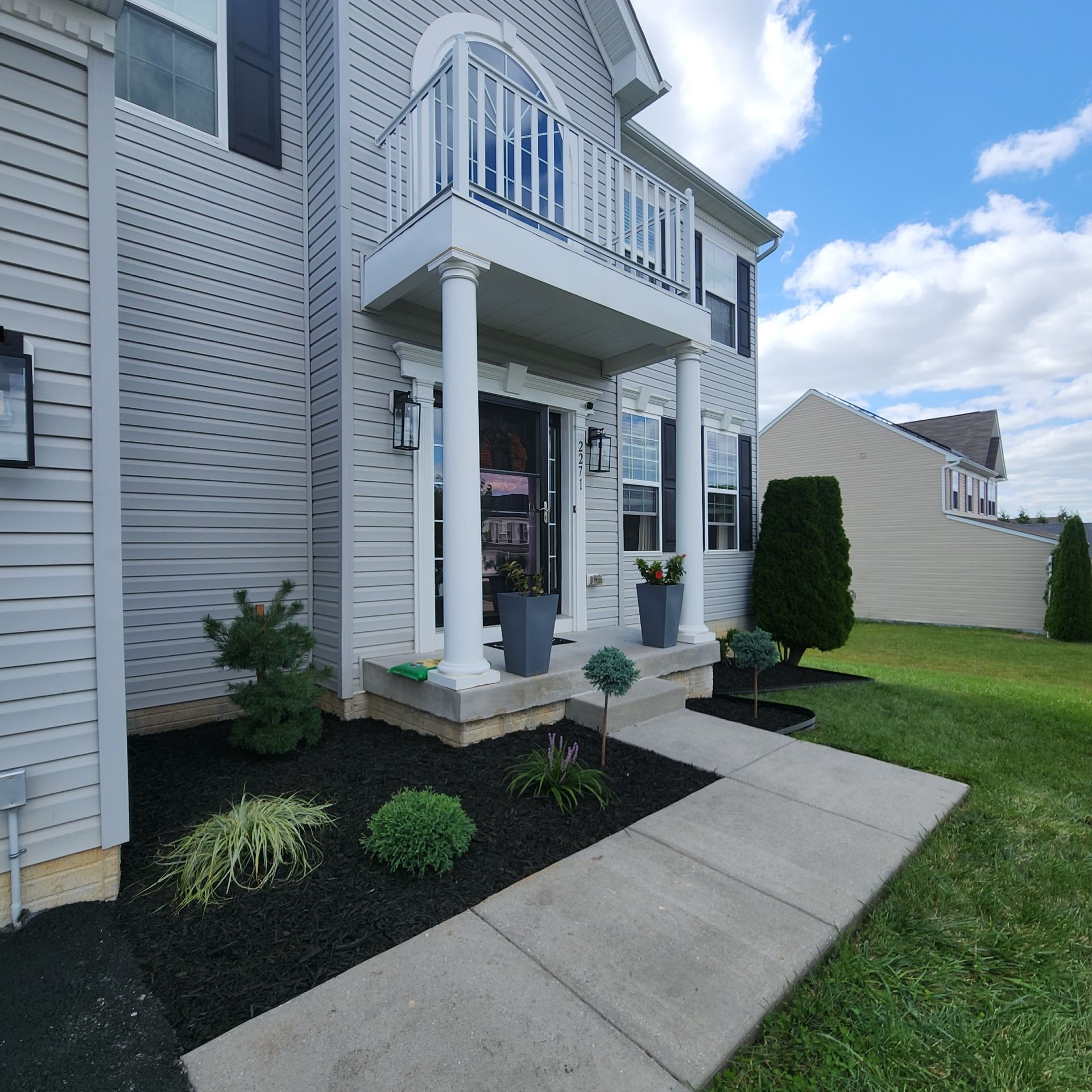 Two-story house with a balcony, pillars, and a walkway surrounded by fresh landscaping under a blue sky.