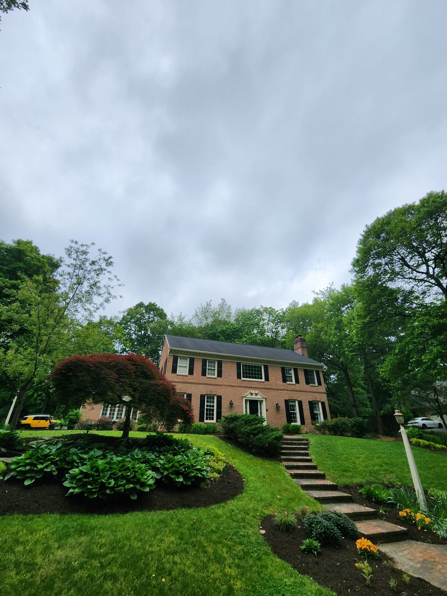 Brick house with black shutters and steps leading to the front door. Landscaped yard, cloudy sky.
