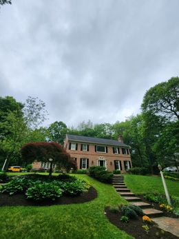 Brick house with black shutters, on a green lawn with a garden and stone steps, under a cloudy sky.