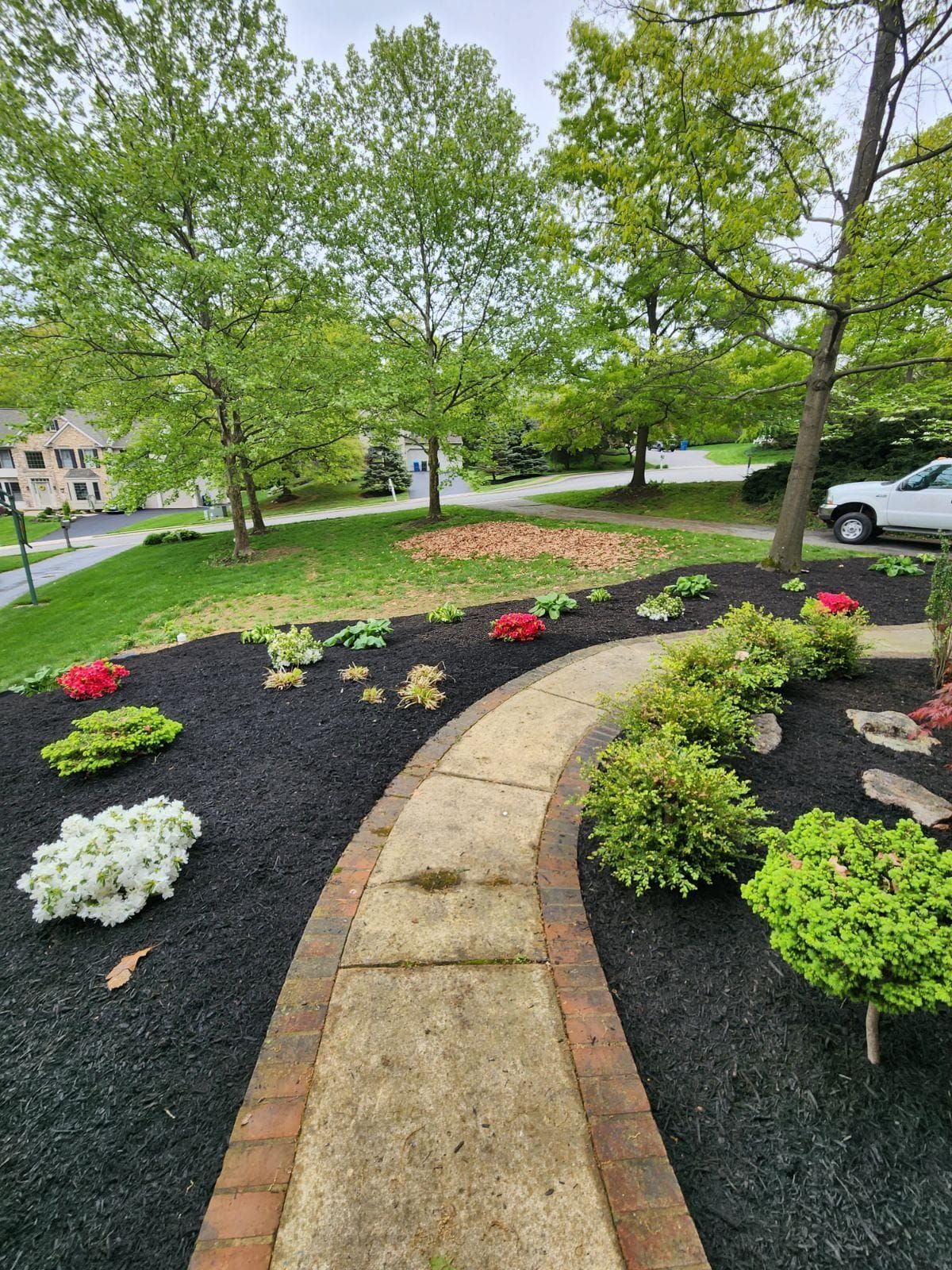 A stone path leads through a garden with azaleas, shrubs, and black mulch. Trees and a distant road in background.