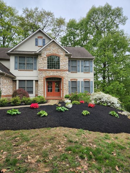 Two-story house with stone facade, red door, and flower beds with dark mulch. Blue shutters, trees in background.
