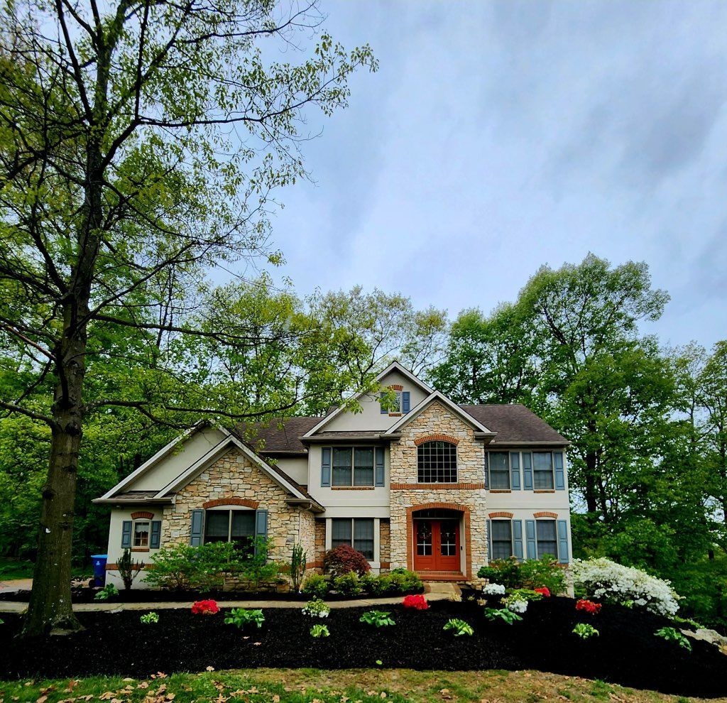 Two-story house with stone facade, red door, blue shutters, and flowerbeds. Trees in the background.