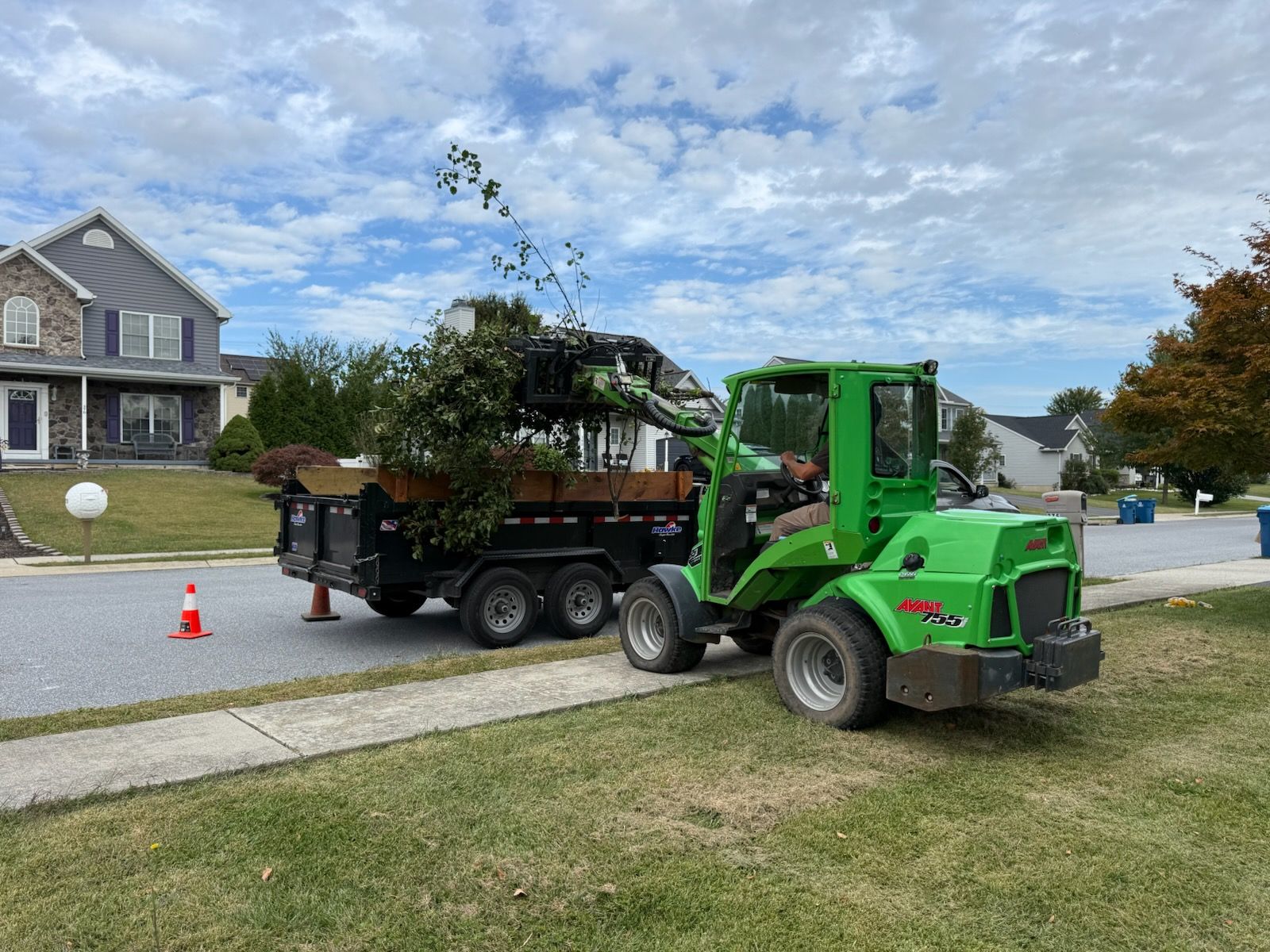 Green loader placing tree debris into a trailer parked on a suburban street.