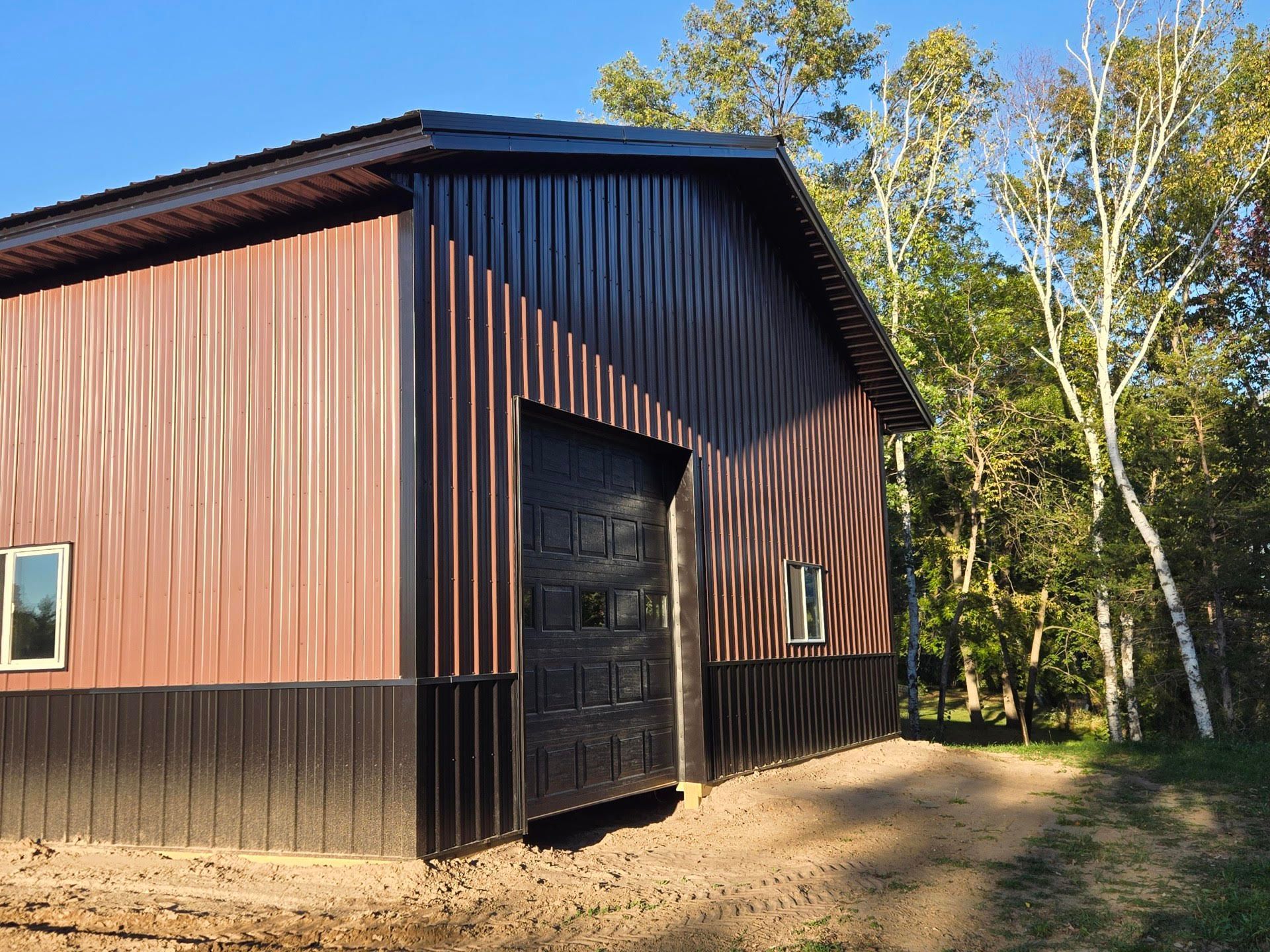 A brown and black barn with a black garage door is surrounded by trees.
