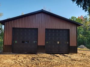 A brown garage with two black garage doors is sitting in the middle of a dirt field.