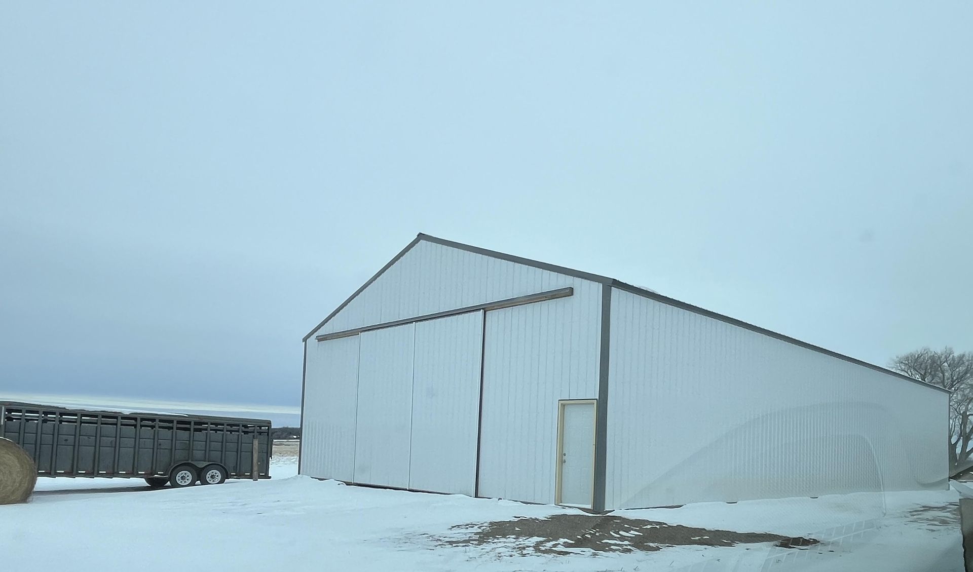 A white barn with a trailer parked in front of it in the snow.
