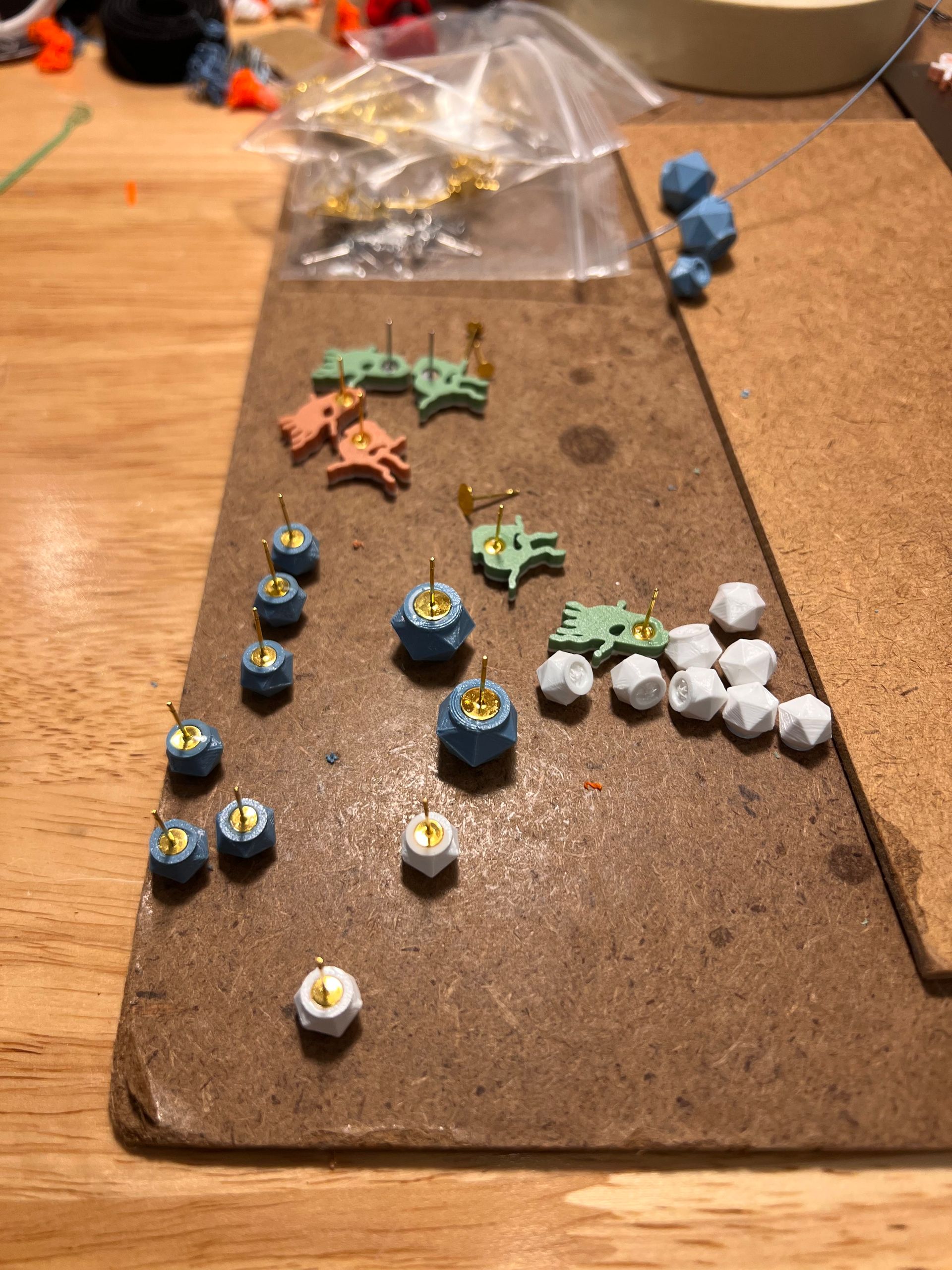 Assorted custom 3D printed earrings fresh from our Brooklyn NY studio. This workbench flat lay features geometric D20 dice studs, playful octopus shapes, and other fun designs in blue, mint green, peach, and white — all printed in durable filament with gold posts, showing the creative and colorful side of our local 3D printing service.
