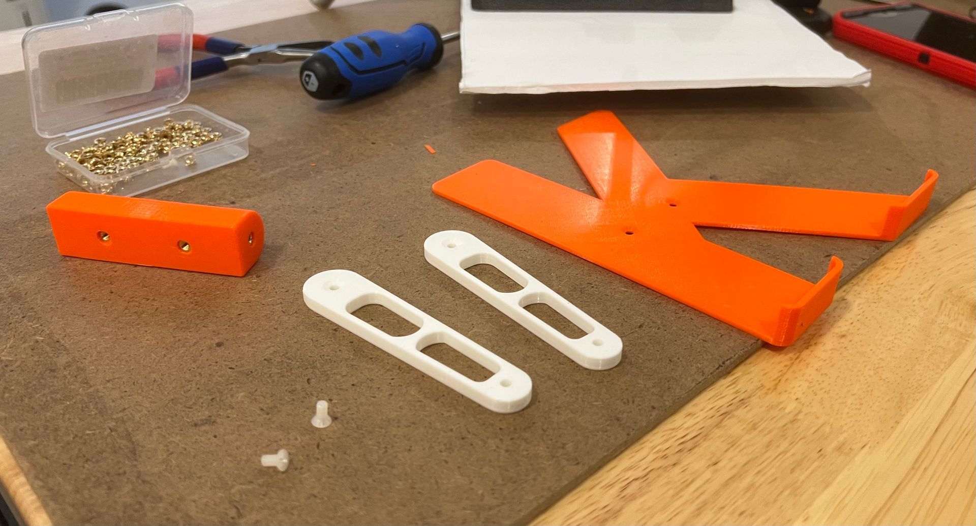 3D printed parts including an orange stand, two white links, a small orange block, and hardware on a workbench.