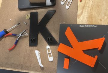 Custom 3D printed letters fresh off the printer in our Brooklyn NY studio. Large black and bright orange “K” shapes sit on the workbench alongside small white brackets, tools, and a Prusa Core One build plate — a real look at how we create bold, durable custom signage and functional parts through our local 3D printing service in Brooklyn.