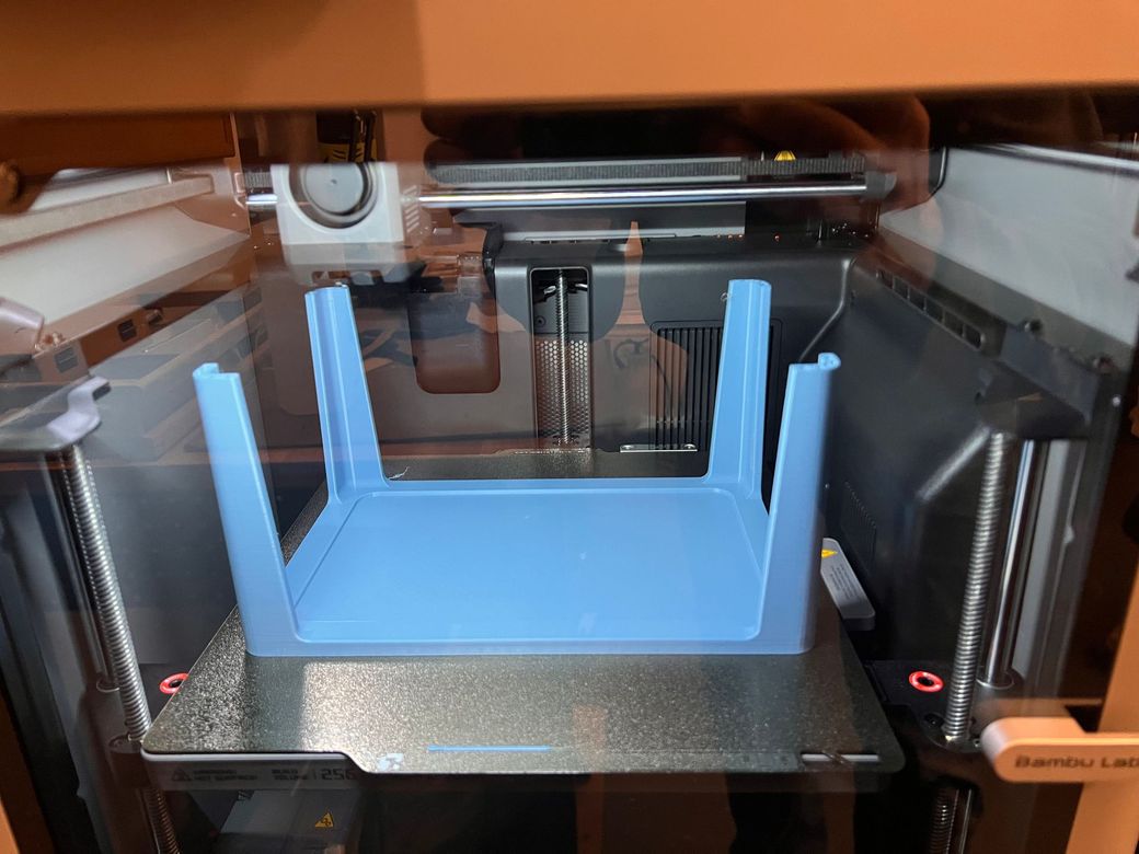 A light blue plastic tray with four legs stands upright inside a 3D printer enclosure.