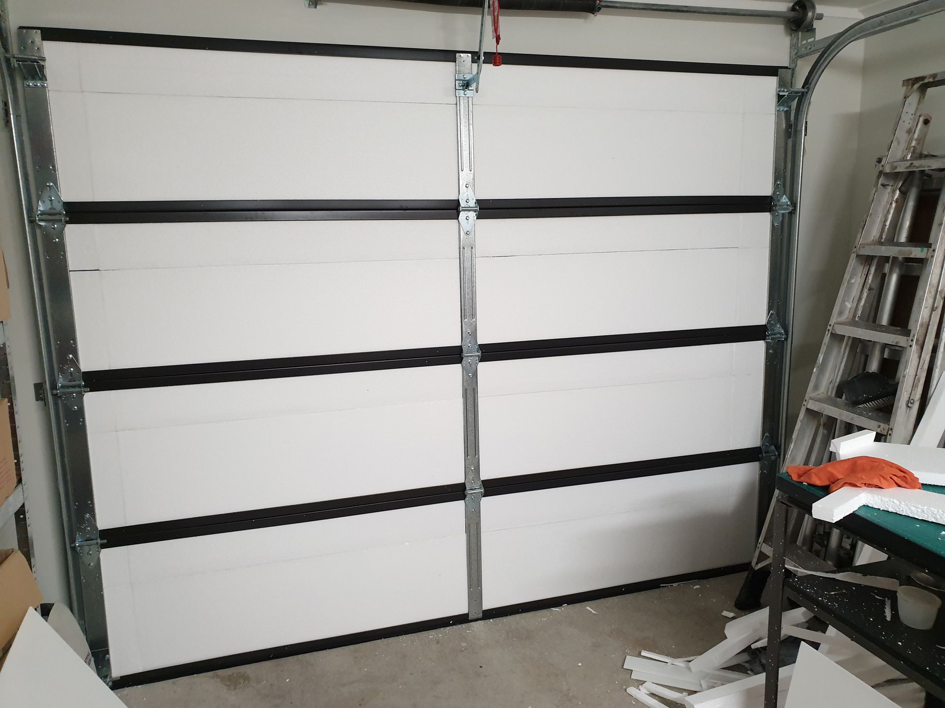 A white garage door with black stripes is sitting in a garage next to a ladder.