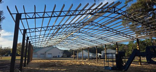 The steel framework and roof trusses of an open-air building under construction on a sunny, grassy lot.