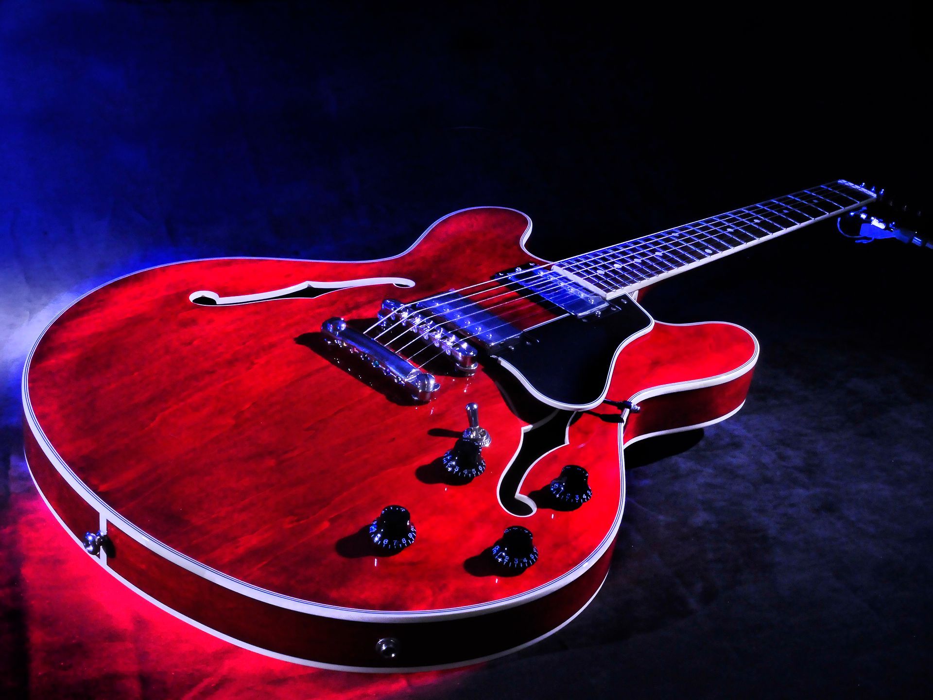 Red electric guitar with silver hardware, illuminated by blue and red light against a dark background.