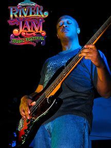Man playing bass guitar at the River Jam Music Festival. Stage lighting, black background, close-up.