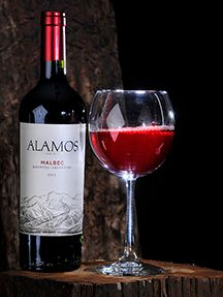 Bottle and glass of Alamos Malbec red wine on a wooden surface against a dark background.