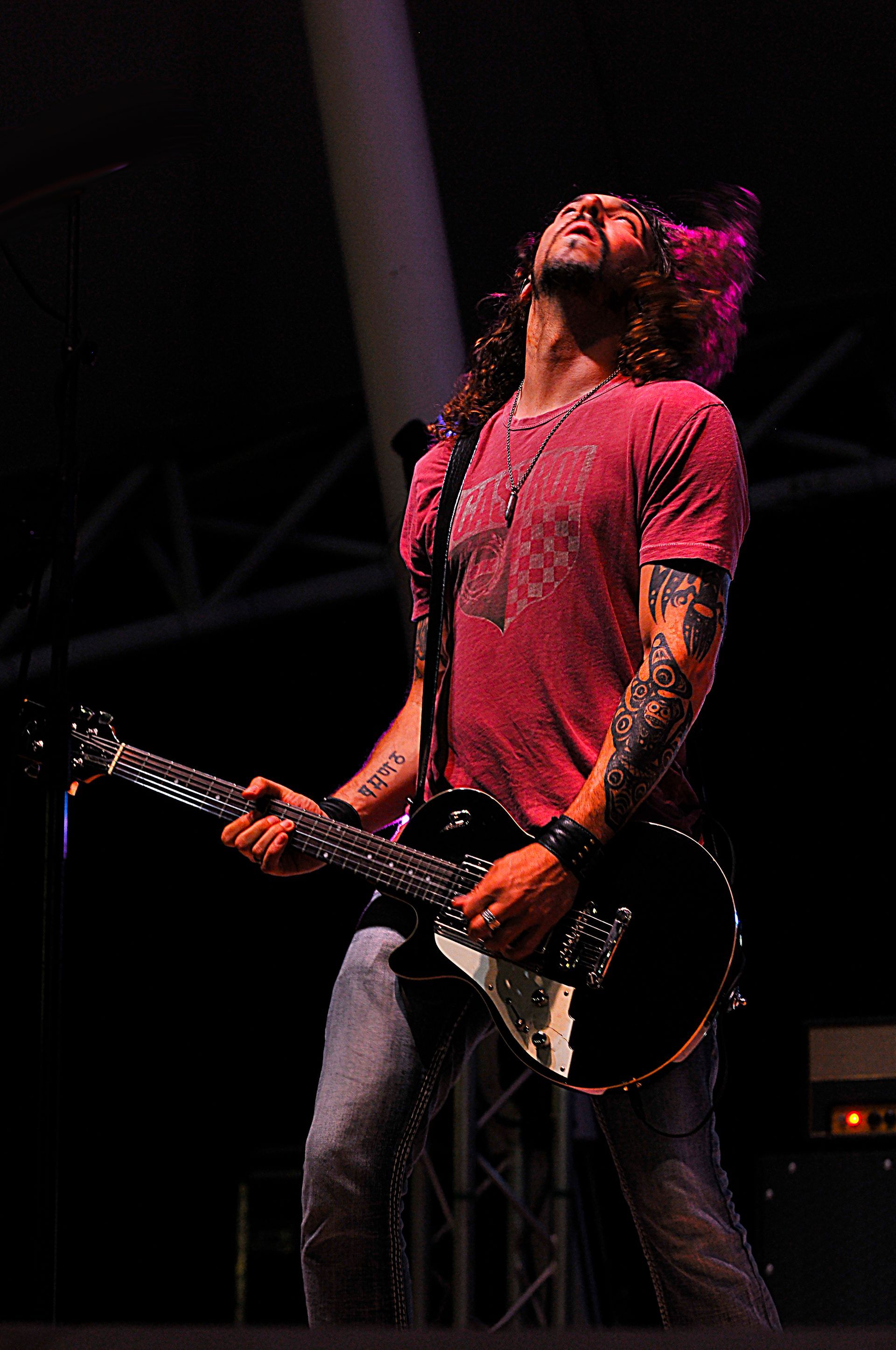 Guitarist on stage, playing a black electric guitar with his head tilted back. He wears a red shirt and jeans.