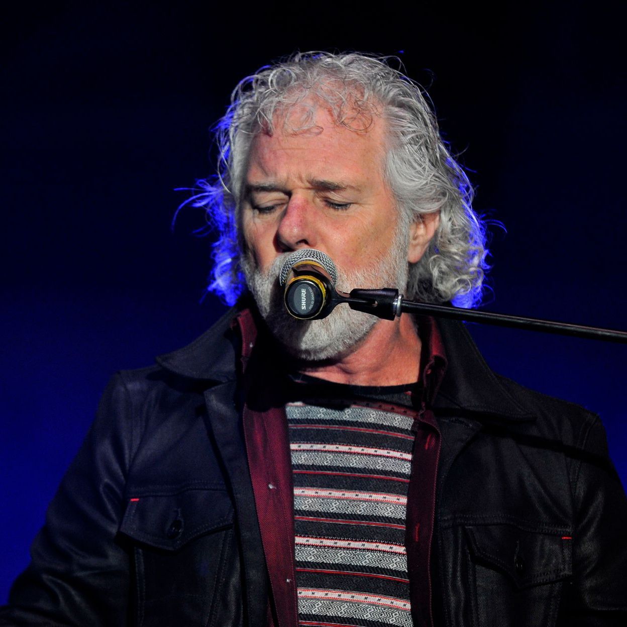 Man singing into a microphone, eyes closed, with a white beard and gray hair, stage lighting.