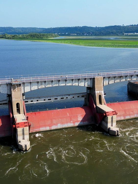 Dam with concrete towers and red gates, holding back a wide river, with land in the background.