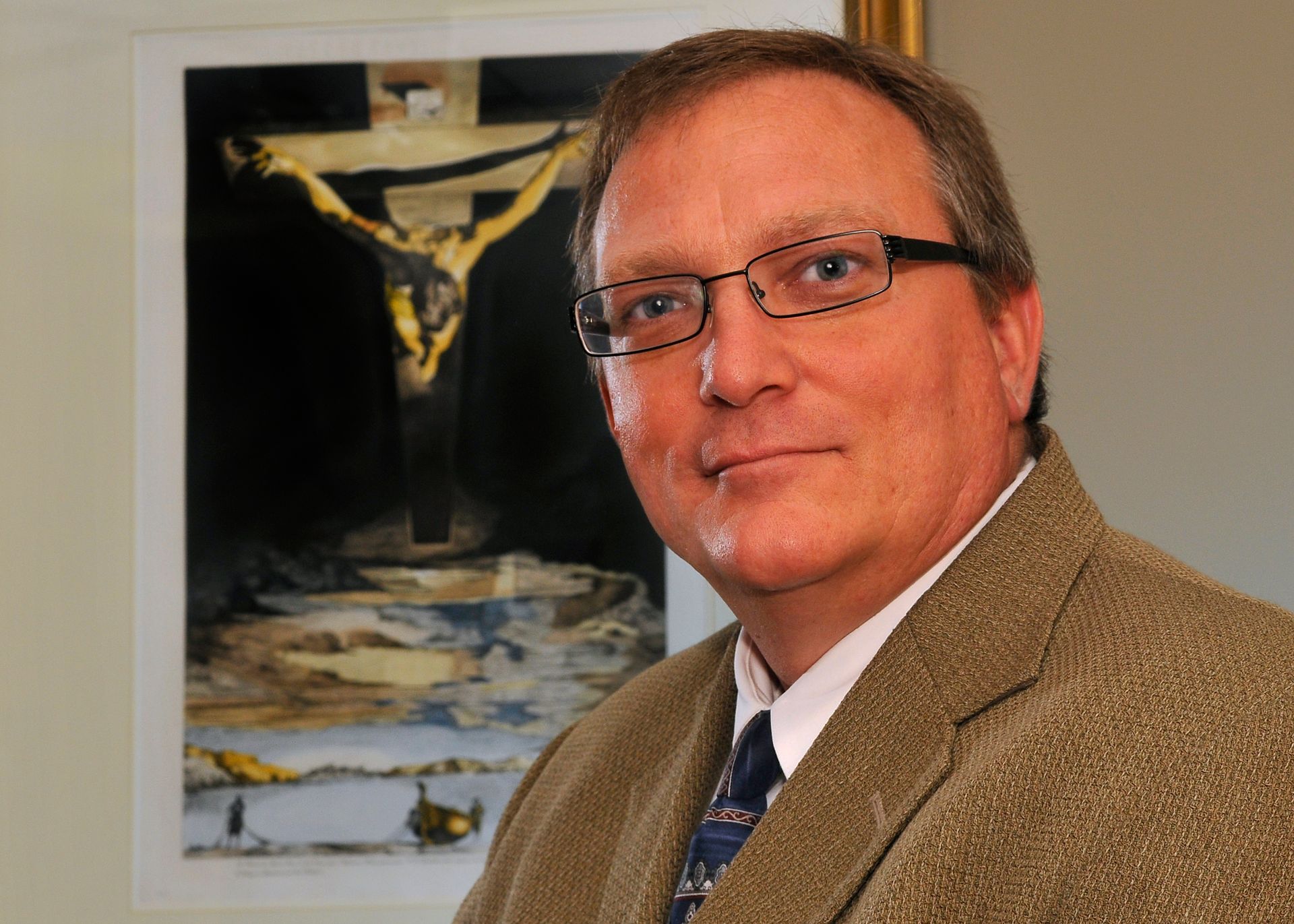 Man wearing glasses and a blazer poses in front of a painting of a crucifixion.