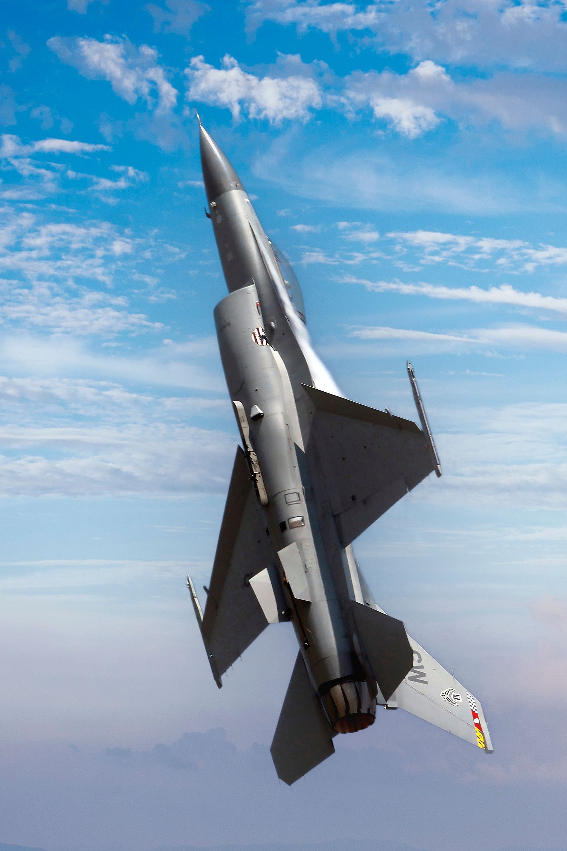 Gray fighter jet banking against a blue sky with scattered clouds.