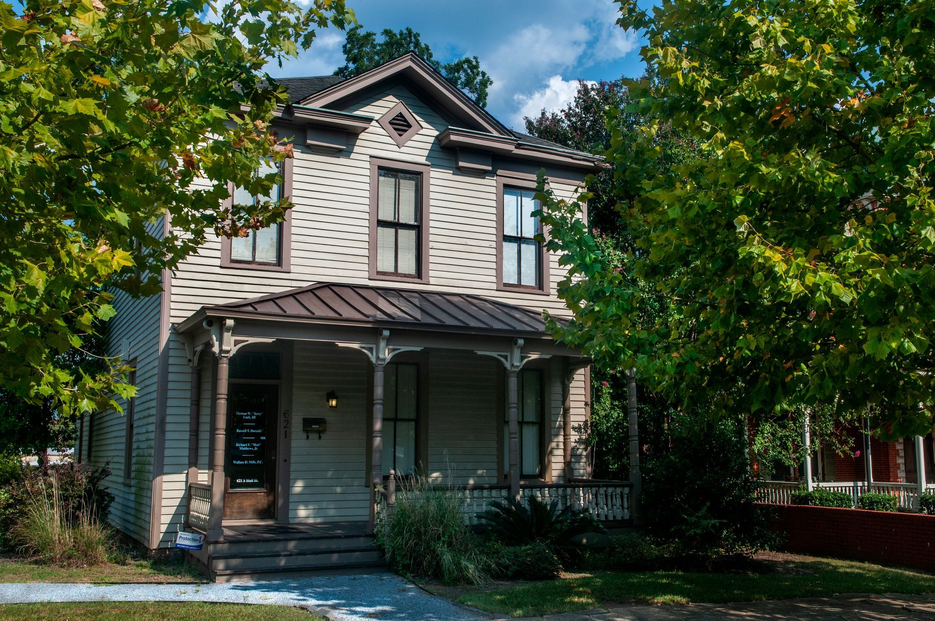 Two-story Victorian house with porch, tan siding, and dark roof, partially obscured by green trees.