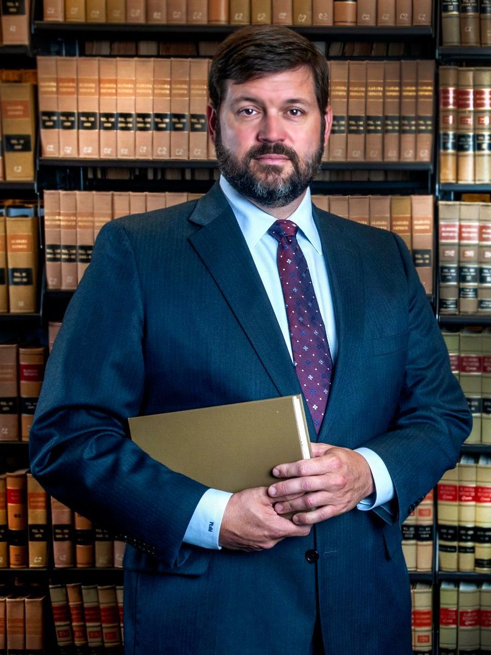 Man in blue suit holding a book, standing in a library with bookshelves.