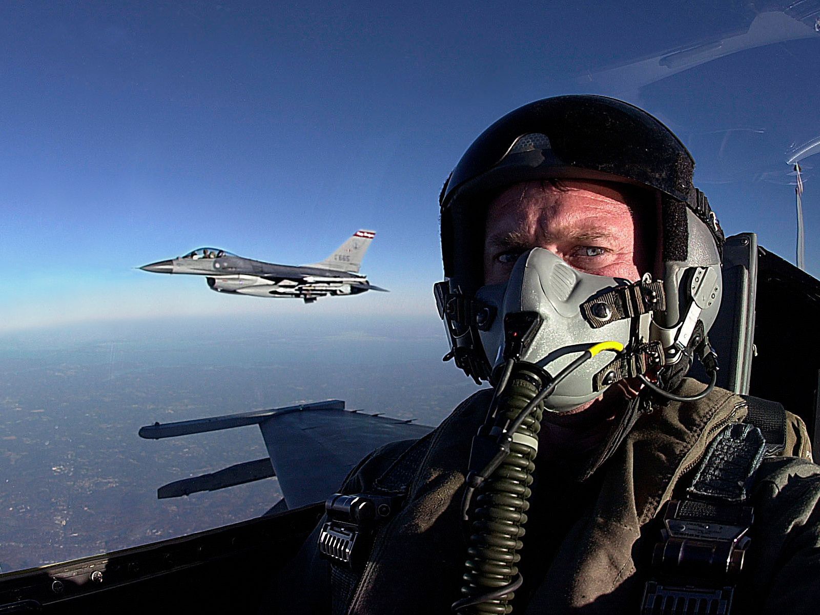 Pilot in cockpit taking selfie with jet fighter in flight, clear blue sky.