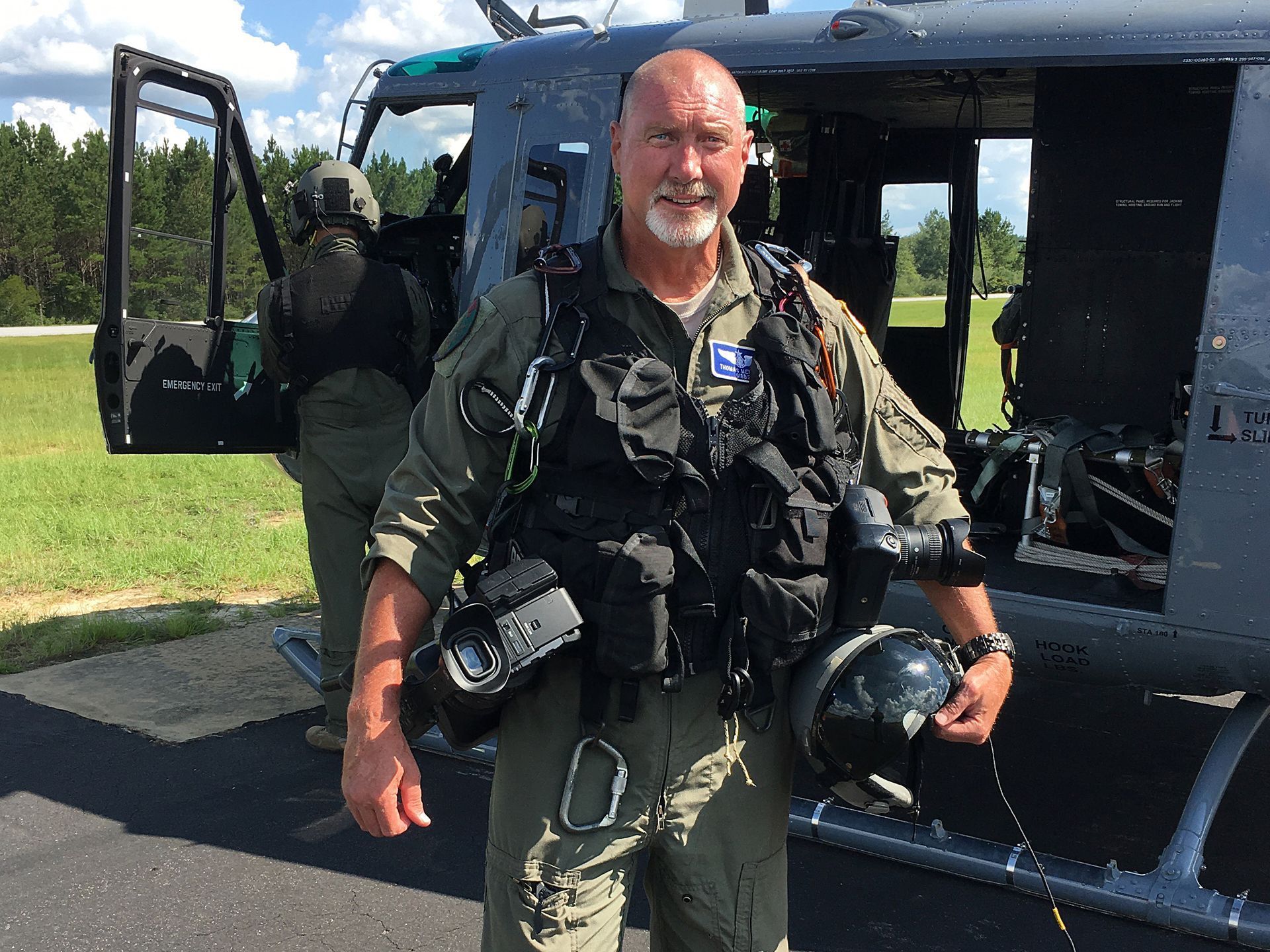 Man in flight gear standing by helicopter, smiling, holding helmet. Another person in background. Outdoors.