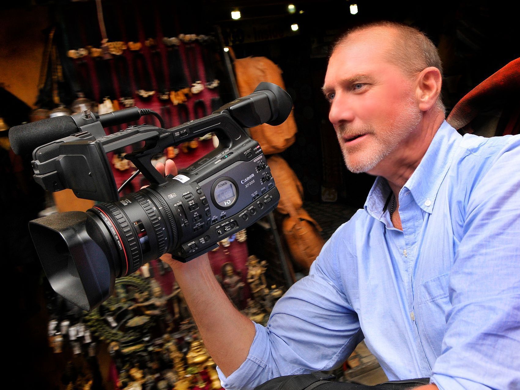 Man holding a video camera, in a blue shirt, filming outdoors.