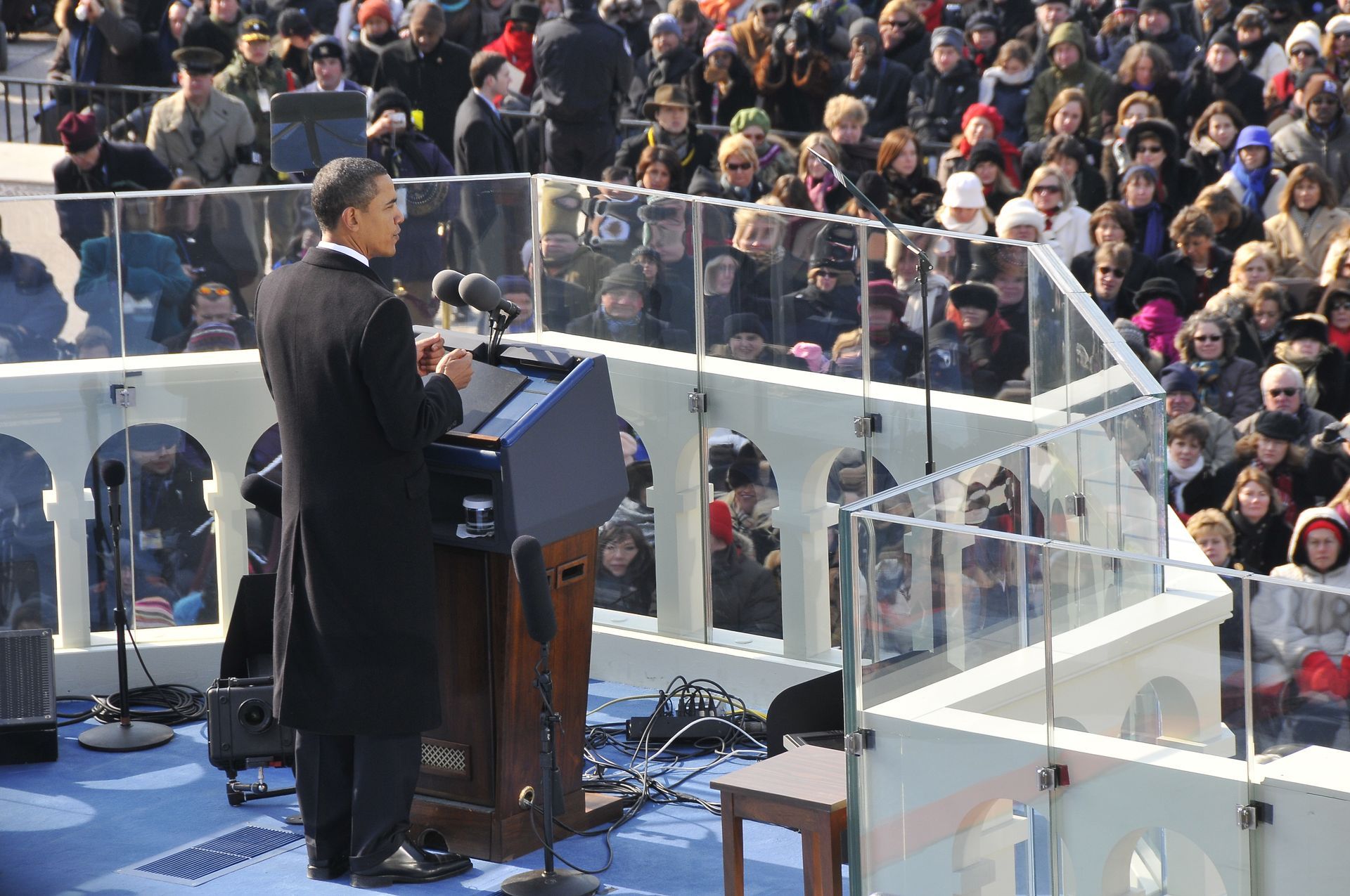 Barack Obama speaking at a podium on an outdoor stage, with a crowd of people in the background.