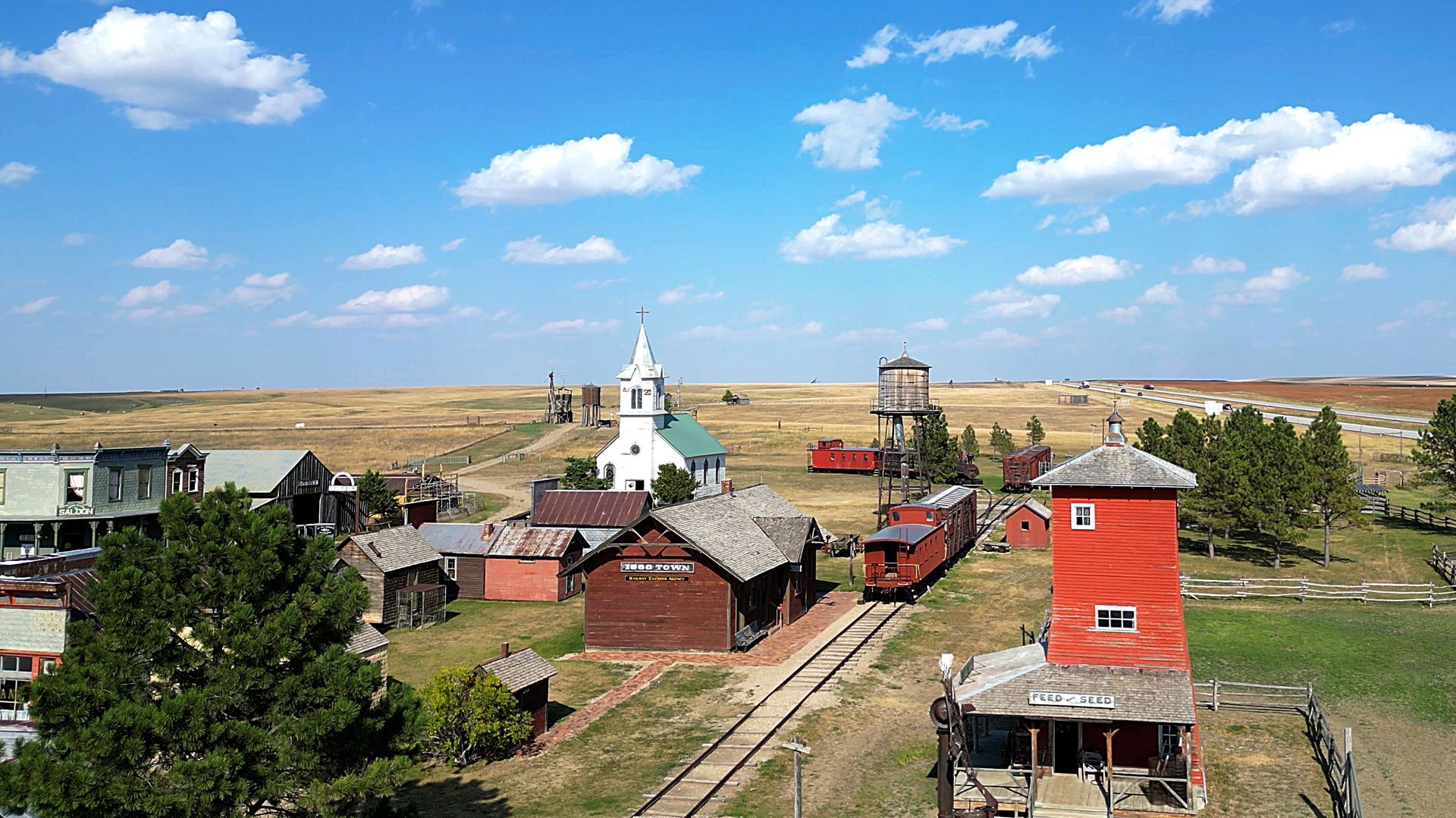 A rustic town with train tracks, a church, and buildings under a blue sky with clouds.