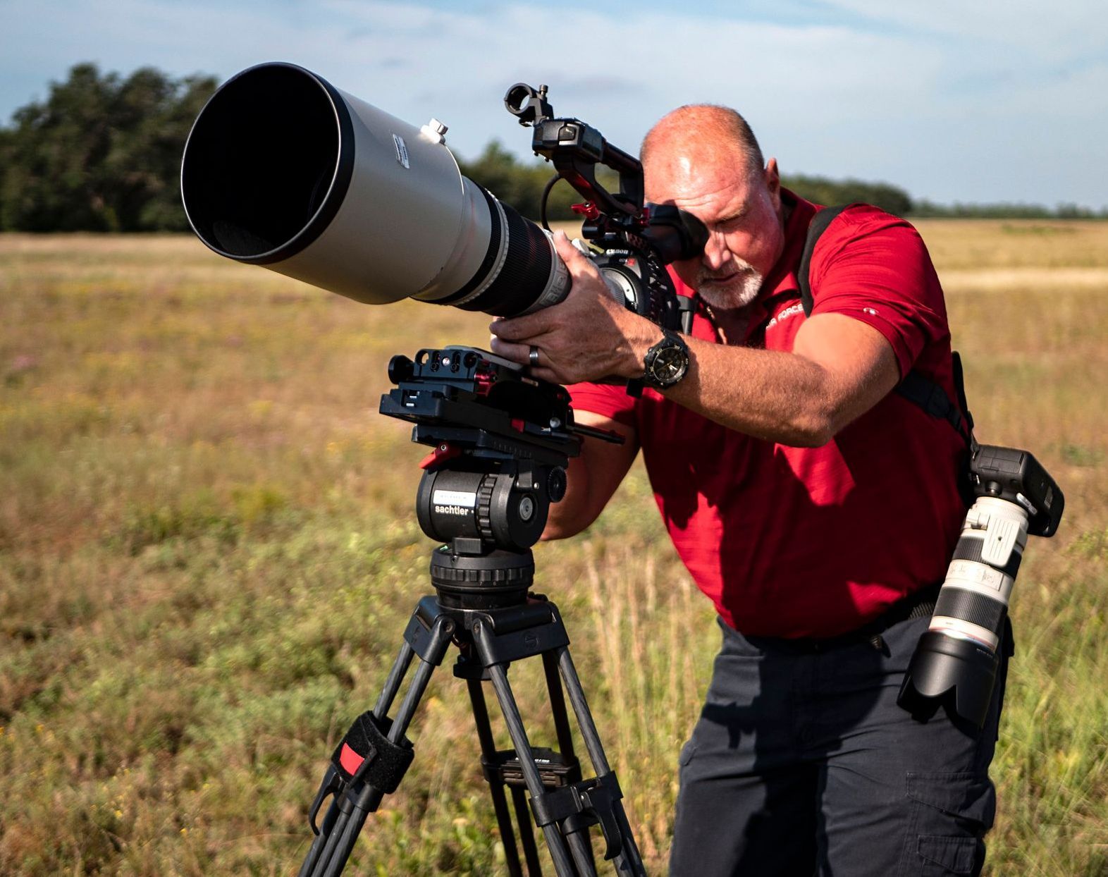 Man using a large telephoto lens on a tripod in a field. He's wearing a red shirt and looking through the viewfinder.