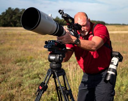 Man using a large telephoto lens on a tripod in a field. He's wearing a red shirt and looking through the viewfinder.