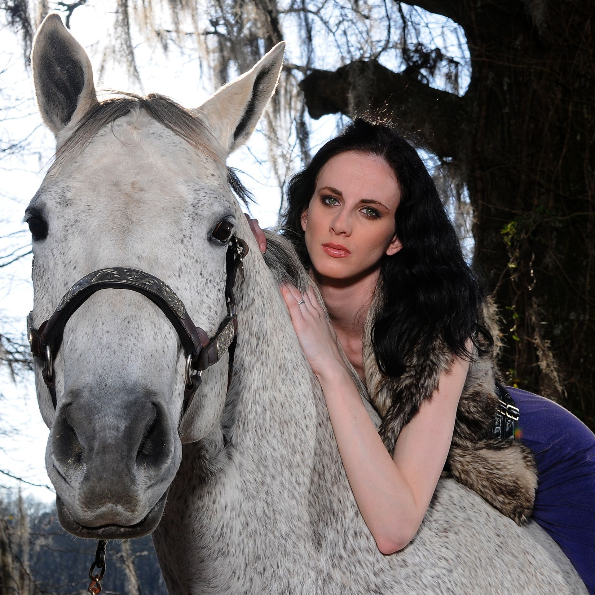 Woman with dark hair, wearing fur vest, on gray horse, looking at camera. Outdoors.