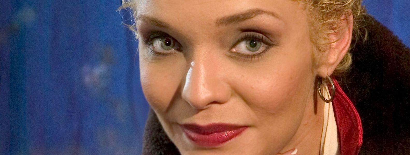 Woman with blonde hair, gray eyes, and red lipstick smiling, wearing a hoop earring. Blue background.