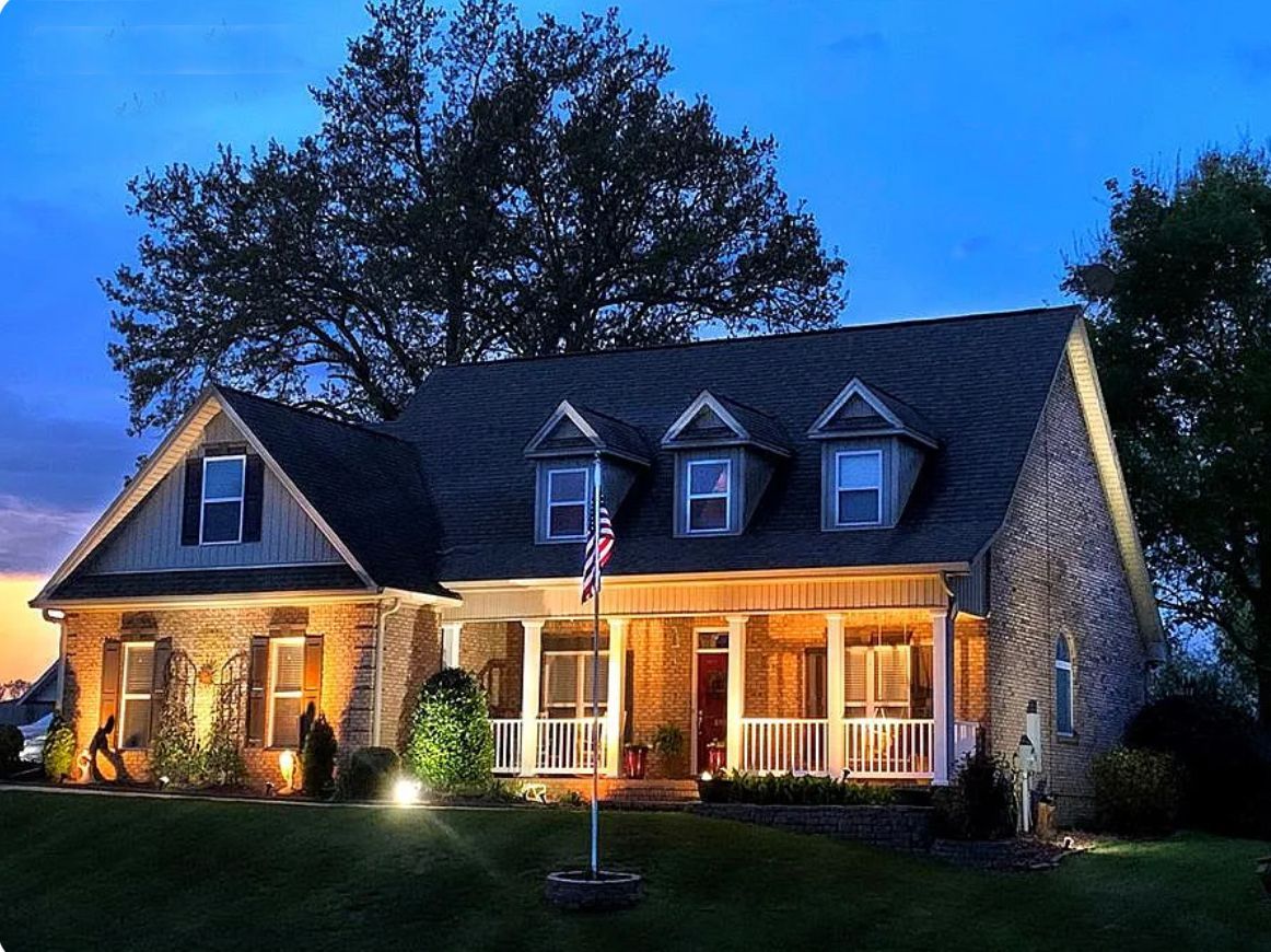 House at dusk, lit by outdoor lights. Stone and wood exterior, porch, and dormer windows.