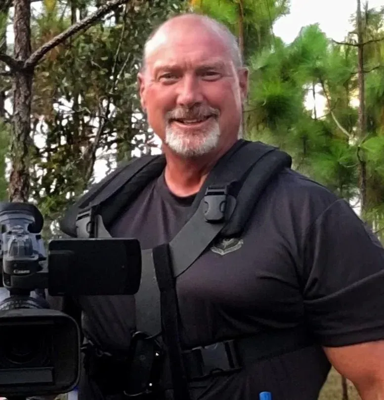 Man in black shirt with camera, smiling outdoors.