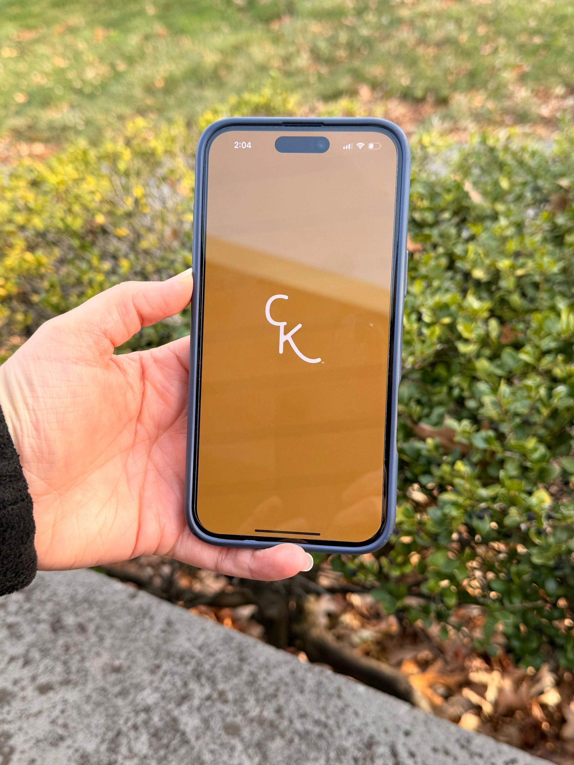 Hand holding a phone displaying a Click it Kitchen against a backdrop of green foliage.