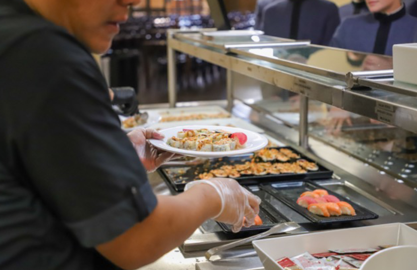 Person preparing sushi rolls behind a buffet counter, holding a plate of food.