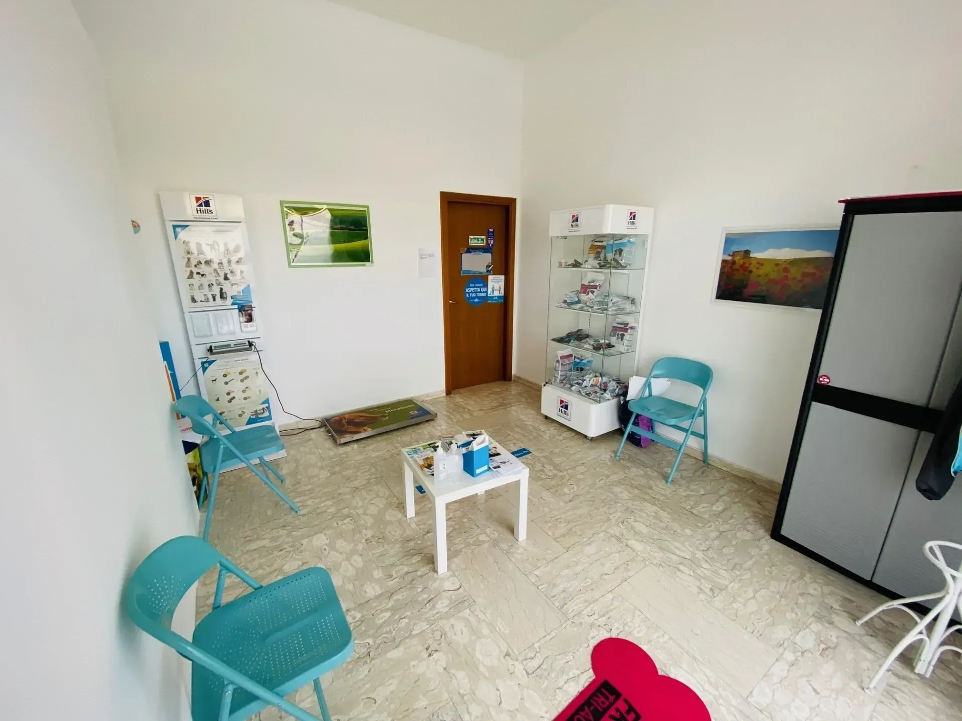 Una sala d'attesa con sedie azzurre, un tavolino bianco, una porta di legno e una vetrina su un pavimento piastrellato.