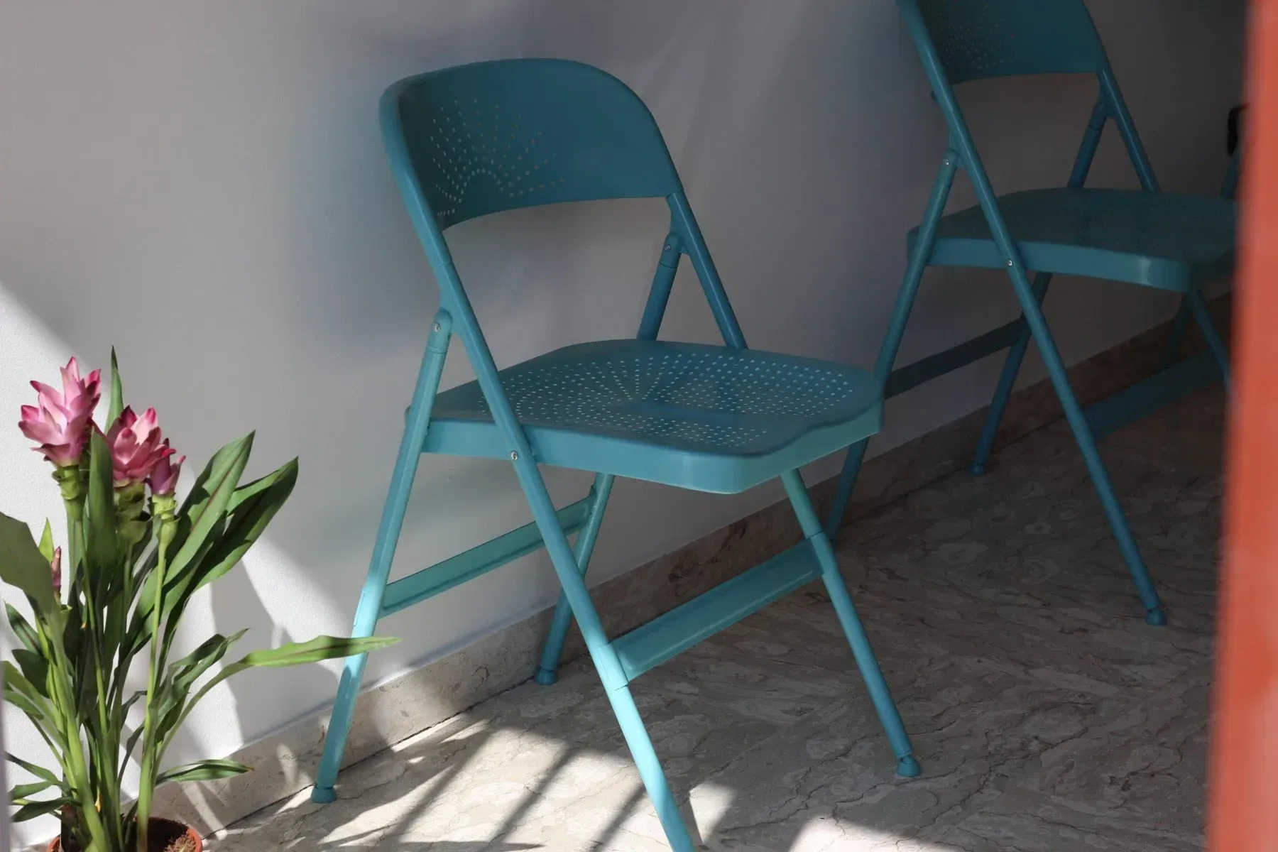 In un angolo soleggiato, accanto a una pianta di fiori rosa in vaso, due sedie pieghevoli in metallo color verde acqua sono posizionate su un pavimento piastrellato.
