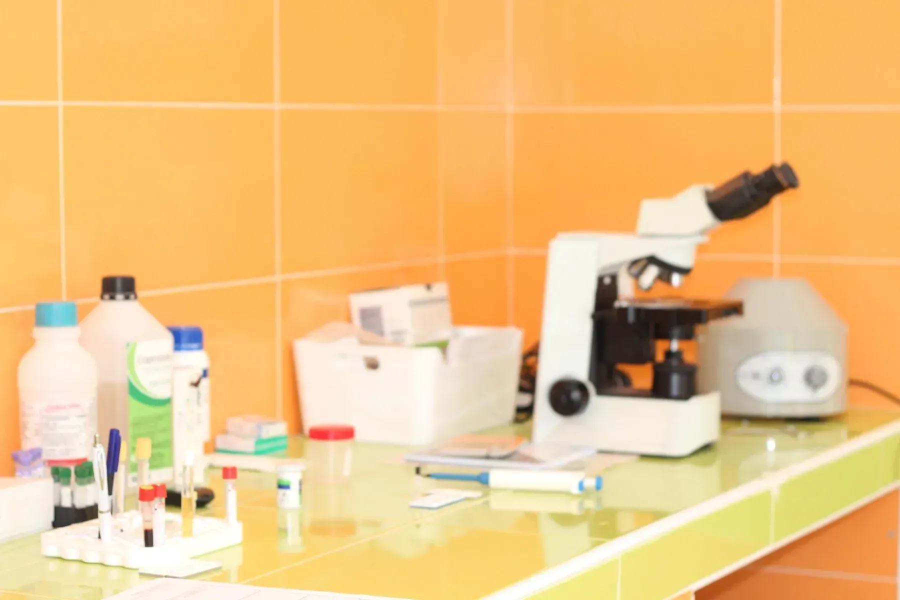 Postazione di lavoro in laboratorio con microscopio, provette e flaconi di prodotti chimici su un bancone addossato a una parete rivestita di piastrelle arancioni.