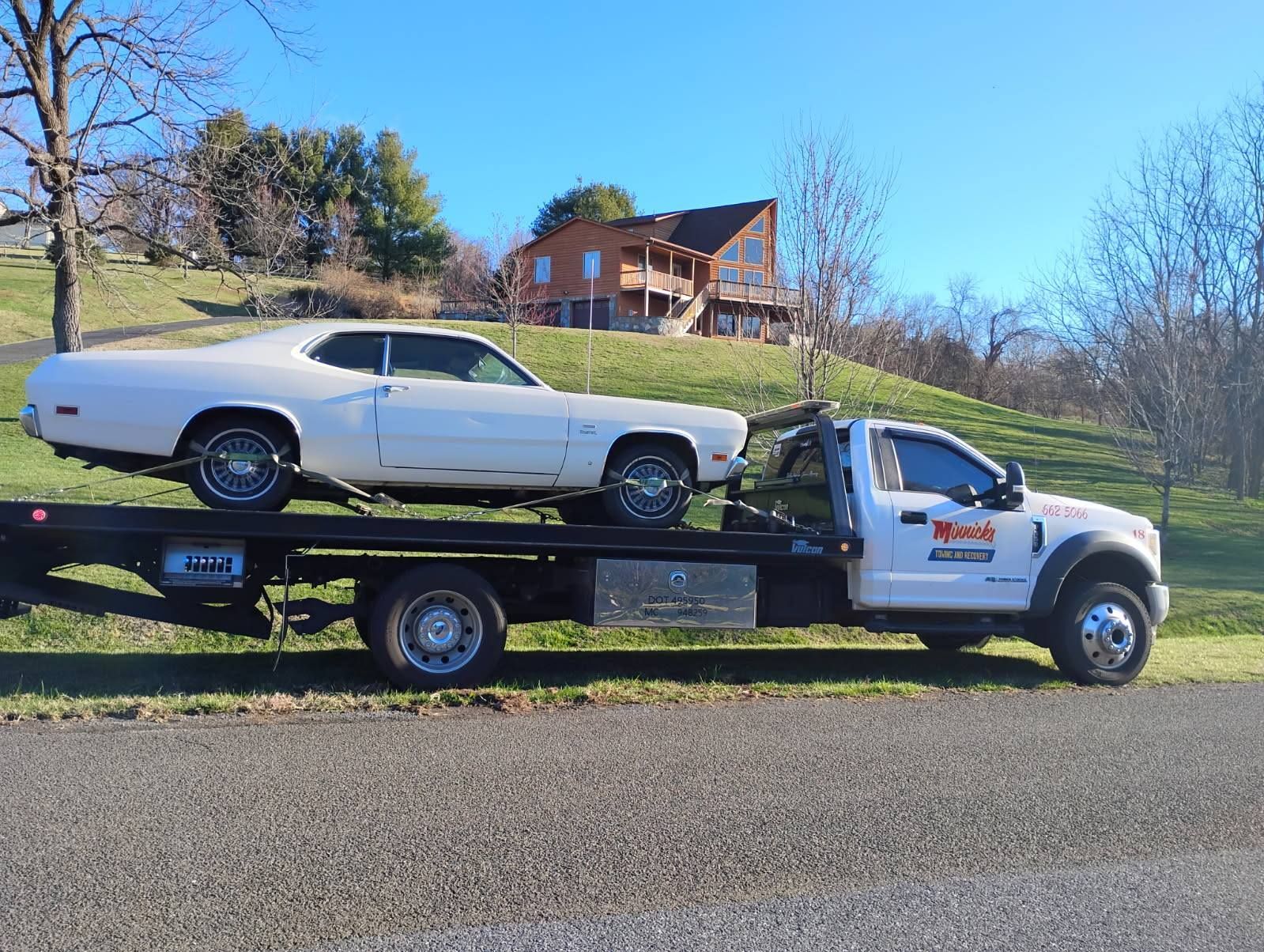 White classic car on a flatbed tow truck. Truck is parked on a road in front of a house.